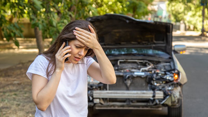 A woman, looking distressed, is on the phone with one hand on her forehead. In the background, a car with its hood open shows visible damage, parked on a leafy street.