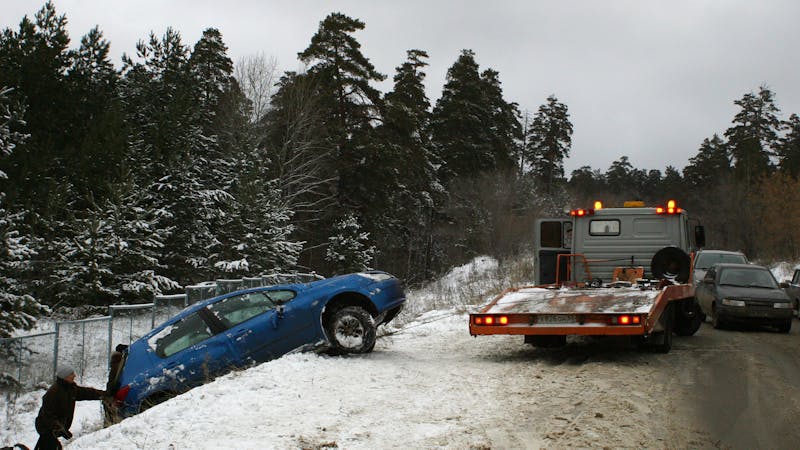 | Matz Injury Law A blue car is partially off a snowy roadside, being pulled by a tow truck. A person is assisting from the ground, while another vehicle waits nearby in a forested area.