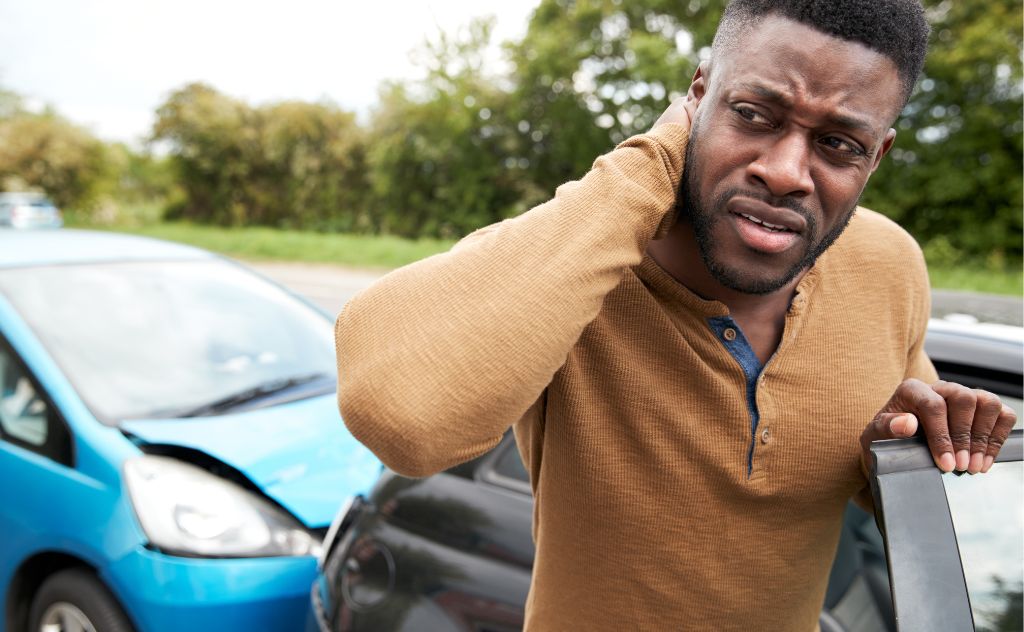 | Matz Injury Law A man stands next to two damaged cars, holding his neck with a concerned expression. He appears to be in distress, in a grassy roadside environment with trees in the background.