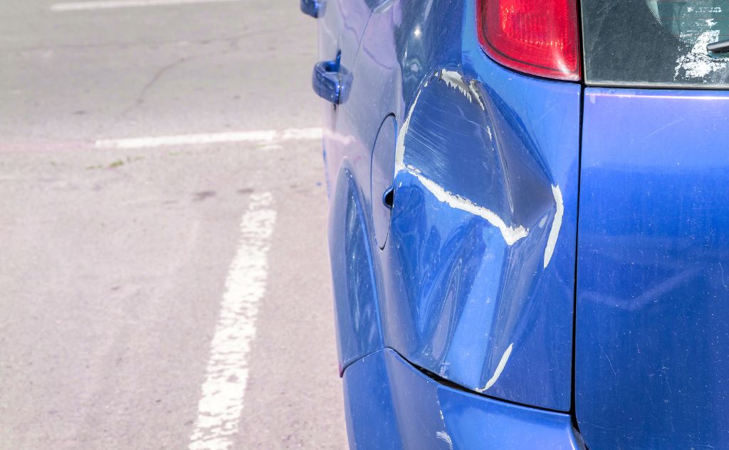 A blue car shows a damaged rear side with dents and scratches, parked on a roadway with faint white lines marking the parking area.