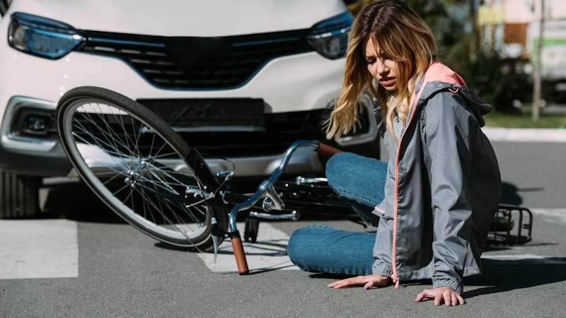 A woman sits on the ground, appearing injured, with a bicycle lying beside her. A man stands by an open car door, looking concerned, on a sunny urban street.