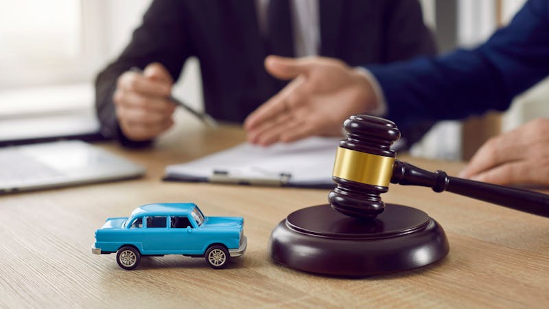 A small blue toy car sits on a wooden table beside a gavel, while two people engage in discussion, suggesting legal or negotiation matters, with paperwork and a laptop in the background.