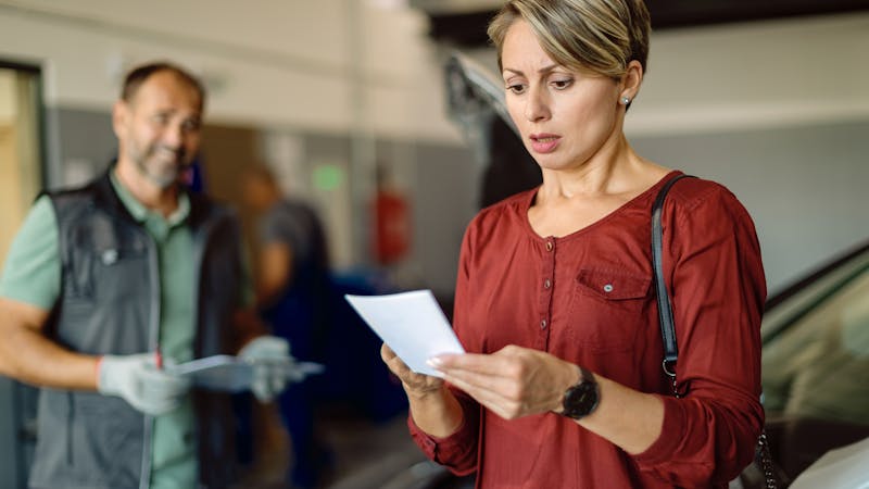 A woman reads a document intently in a garage as a mechanic waits with paperwork, suggesting a car servicing discussion.