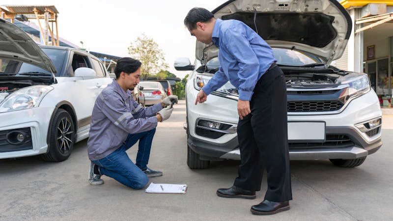 | Matz Injury Law A mechanic kneels by the front of an open car hood, inspecting a component, while a businessman stands beside him, pointing at it. Nearby, two other cars are parked.
