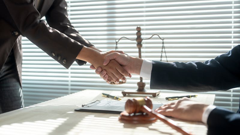 Two hands engage in a handshake across a table. A gavel and legal scales are positioned nearby, with sunlight filtering through horizontal blinds, creating a formal, professional atmosphere.