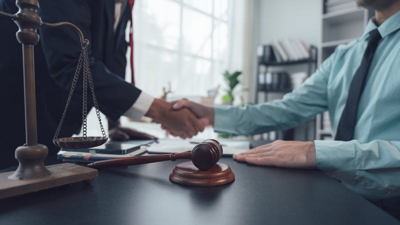 A gavel rests on a desk while two individuals shake hands, signifying agreement or a deal, in a professional office with bookshelves and large windows in the background.