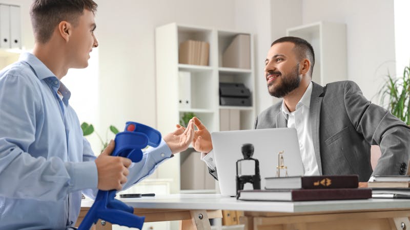 A young man in a blue shirt holds a blue object while engaging in conversation with a man in a gray suit in a modern office setting, featuring bookshelves and plants.