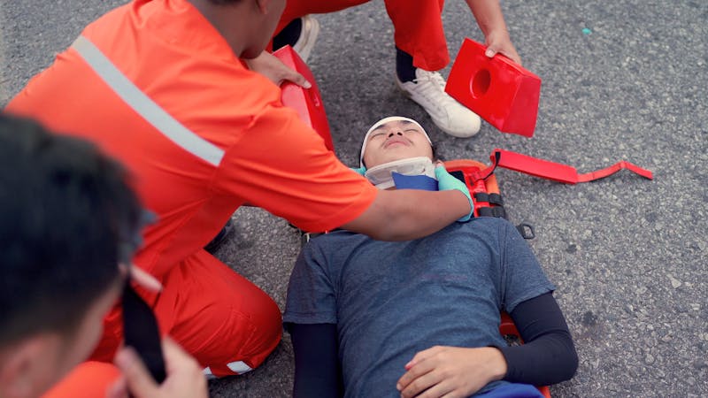 Paramedics attend to an individual lying on the ground, securing a neck brace, in an emergency medical situation outdoors.