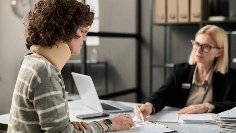 woman wearing a neckbrace talking to another woman at desk. 