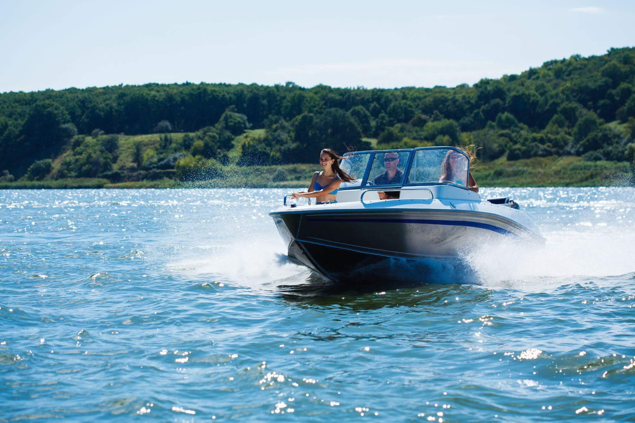 Three people are traveling on boat in a lake, the day is sunny and there's a forest in the background \