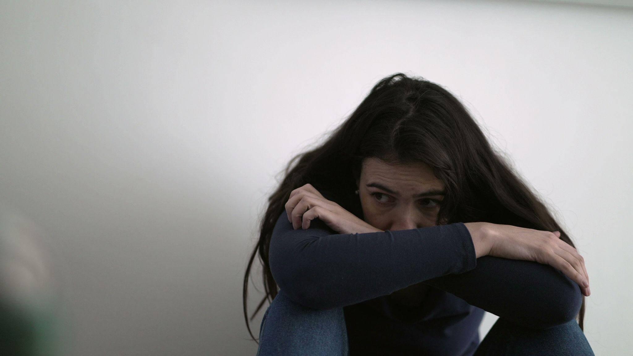 Woman sitting on floor with hands on knees indoors. She seems distressed