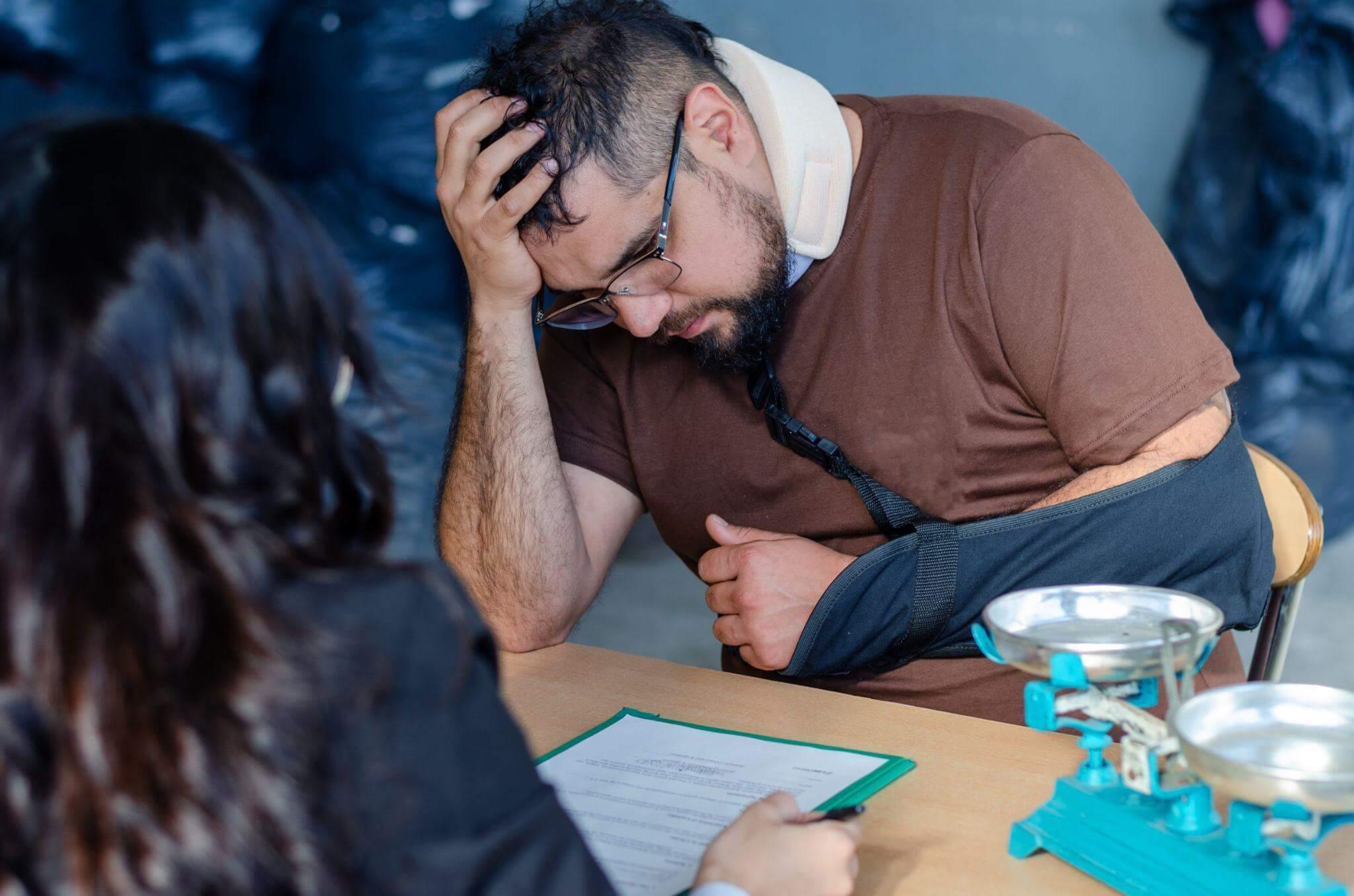 a man sitting at a table in front of a woman, he is wearing an arm and neck brace, he seems worried