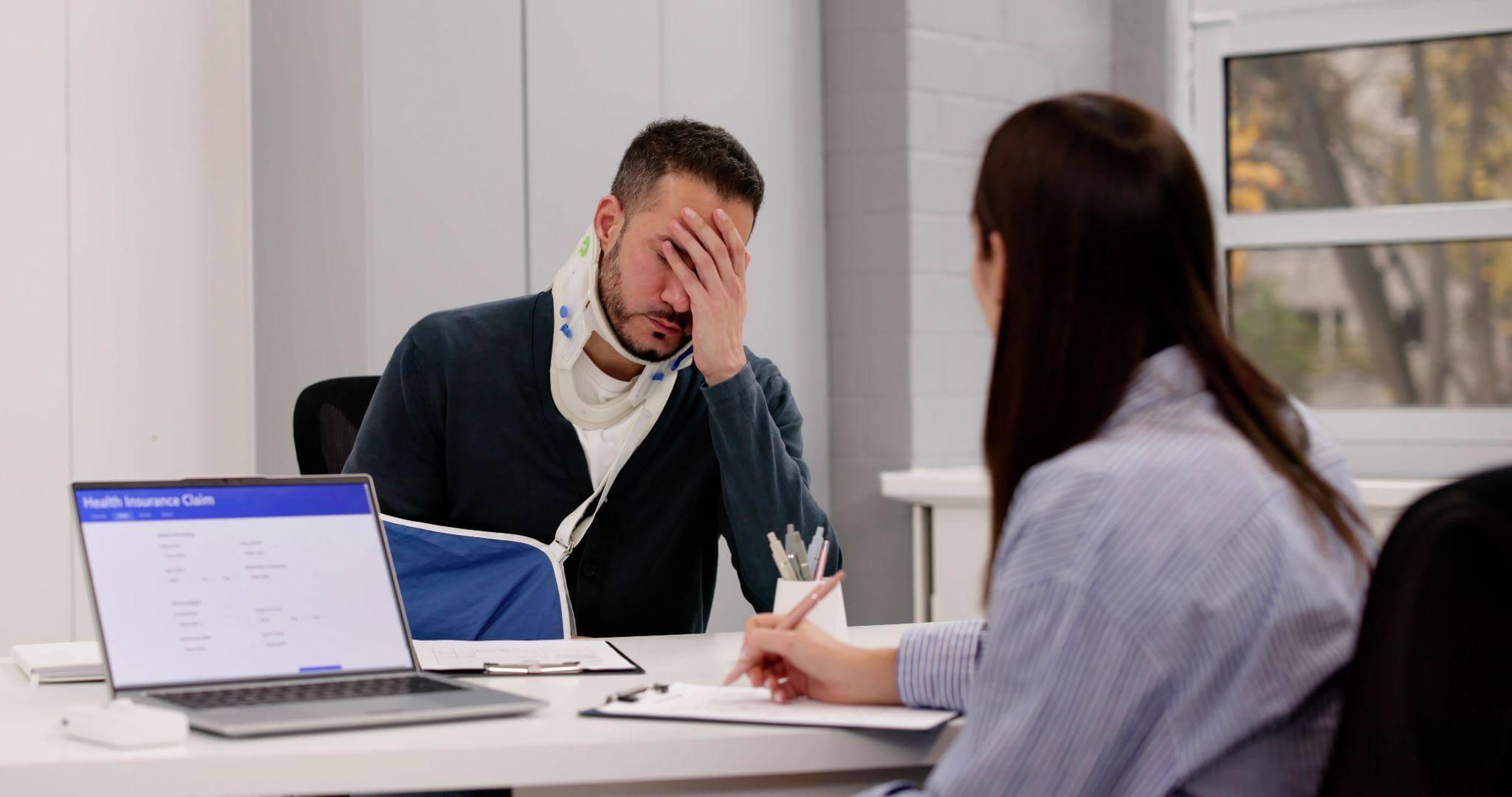 Man and woman discussing at an office desk, the man is wearing a neck and arm brace and seems to be in distress