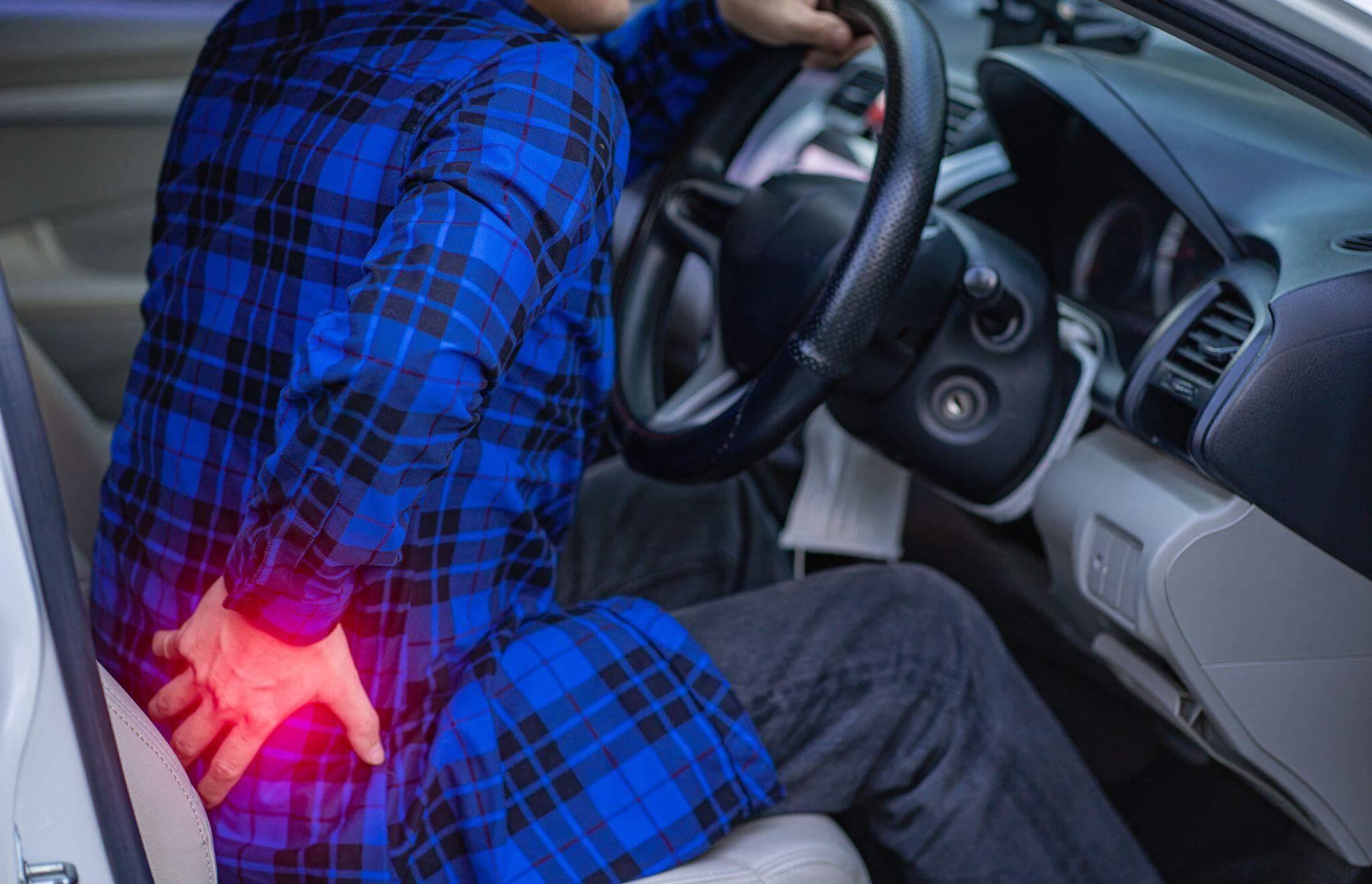 Man driving car holding steering wheel with one hand in touching his back with the other hand indicating acute pain