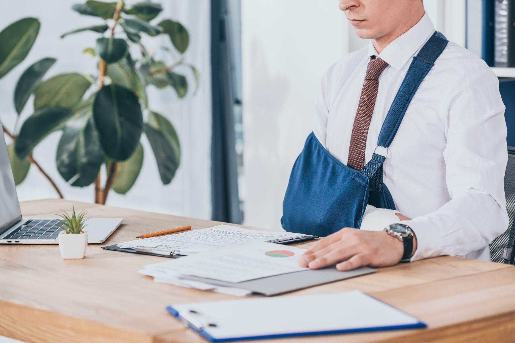 Man working at a desk, there are multiple papers and a laptop, he is wearing an arm brace