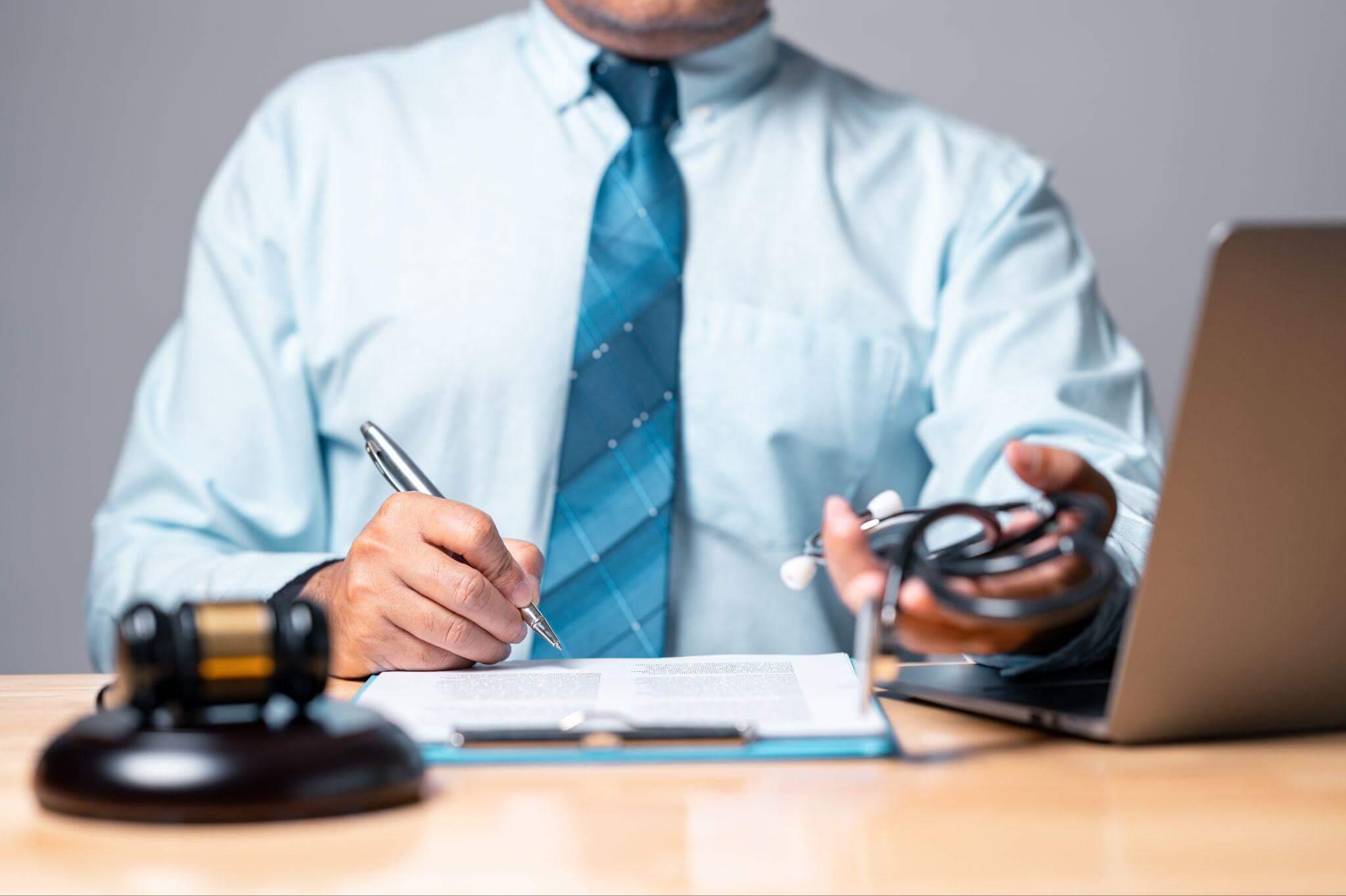 Man writing notes at desk, there's also a gavel and a stethoscope 