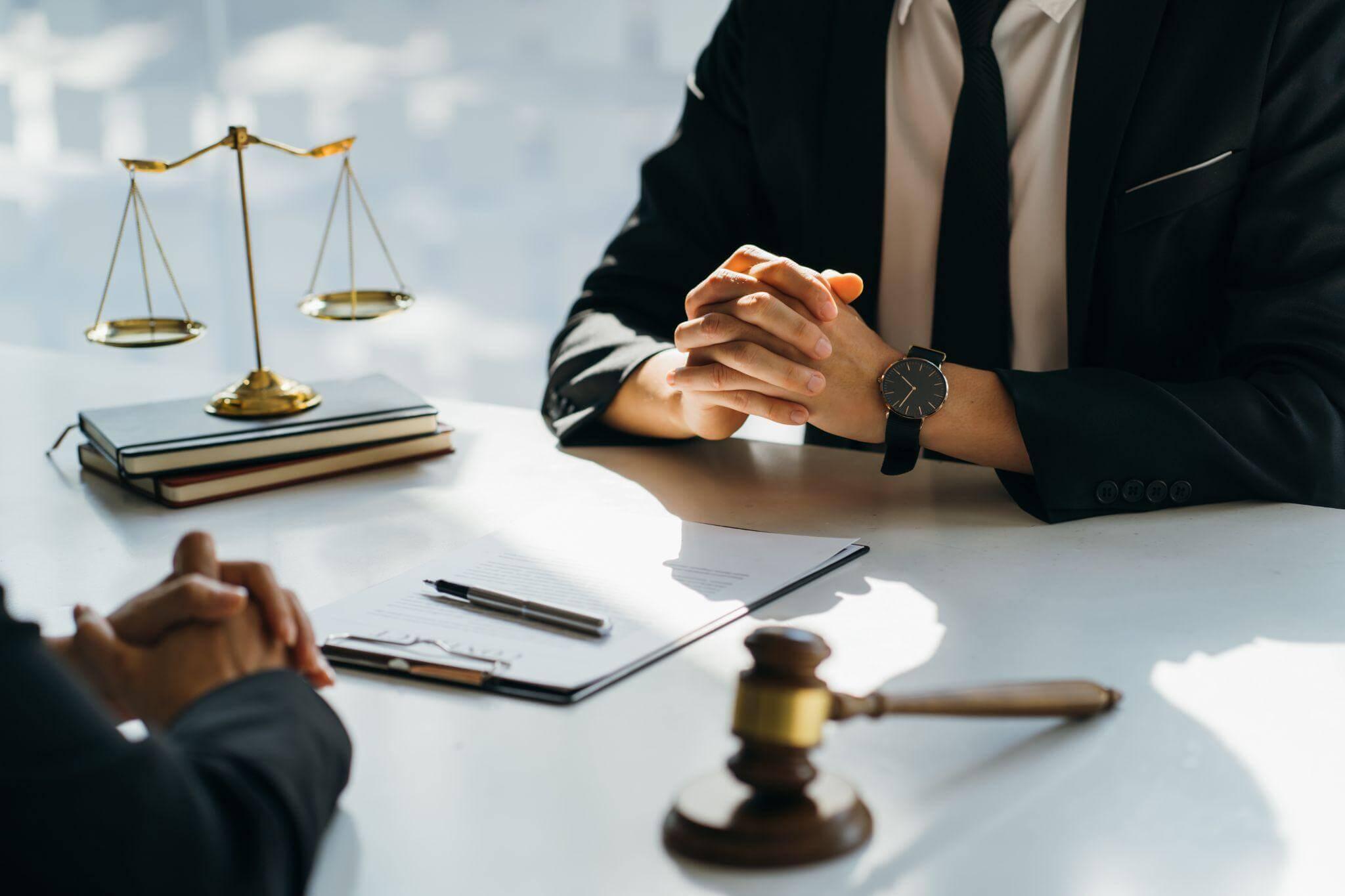 Two people having a discussion at a table, there's a gavel, scales, books and paper on the table