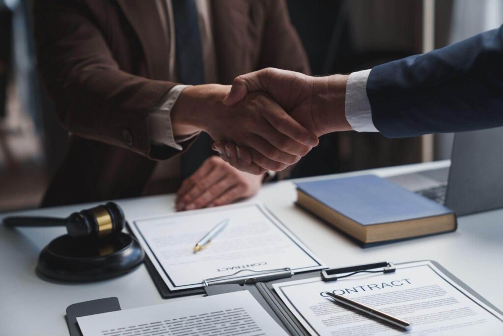 Business professionals shaking hands over signed contracts on a desk.
