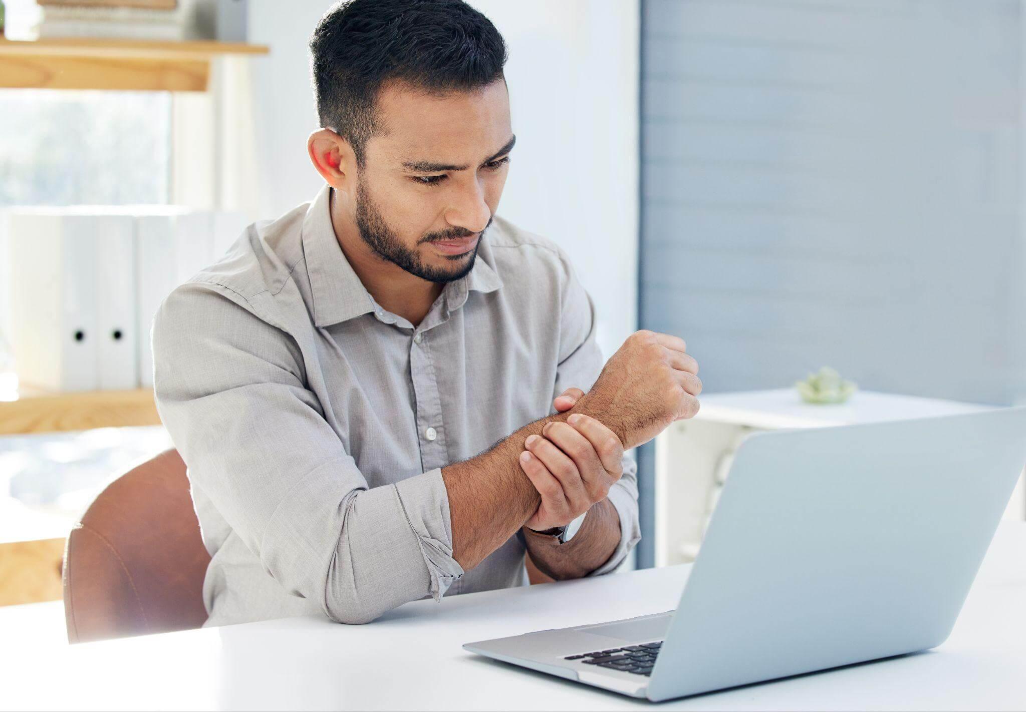 a man sitting in front of a laptop computer, he is touching his wrist and seems to be in pain